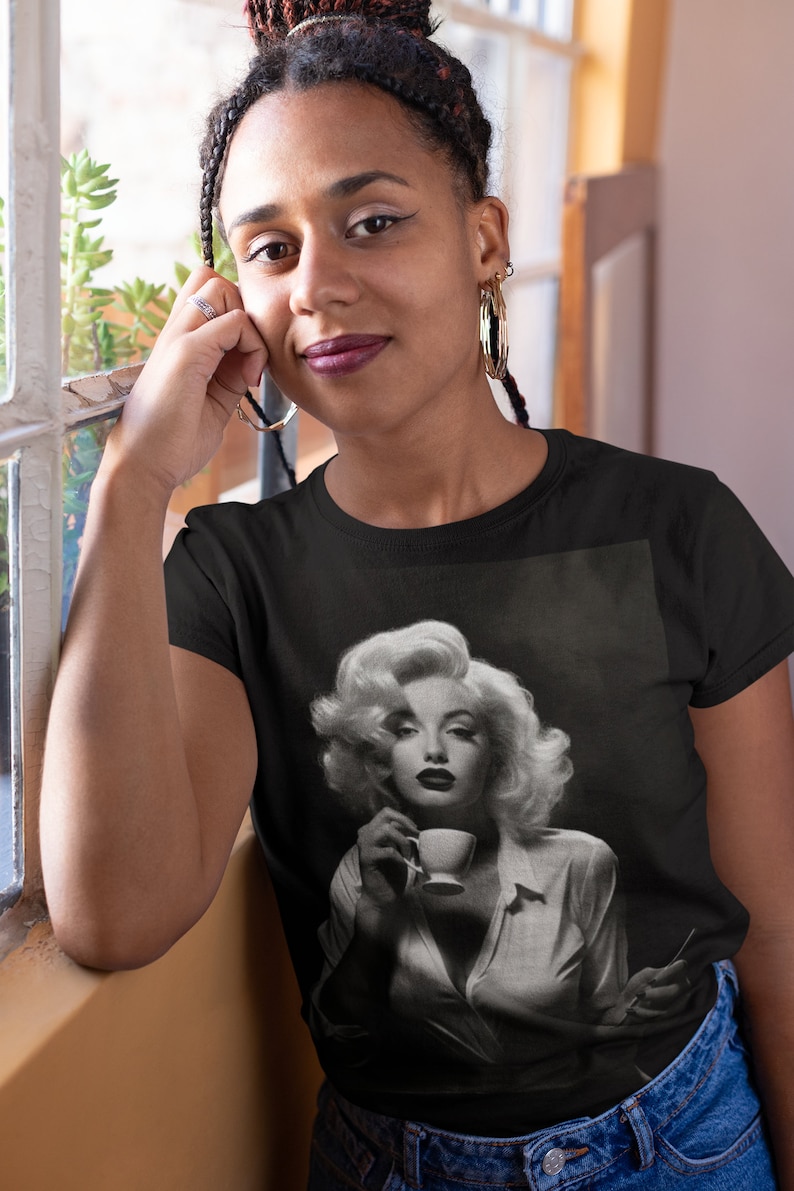 May include: Black t-shirt featuring a black and white image of Marilyn Monroe holding a teacup. The image is a close-up of the actress, with her blonde hair styled in voluminous curls. The shirt is a classic crew neck.
