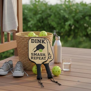 Könnte beinhalten: Zwei Pickleball-Schläger aus Holz mit dem Text "DINK, SMASH, REPEAT AT" werden auf einer Holzterrasse gezeigt. Ein Korb mit gelben Pickleballs, eine Wasserflasche und ein Paar graue Turnschuhe sind ebenfalls zu sehen. Die Szene deutet auf Freizeitaktivitäten im Freien hin.