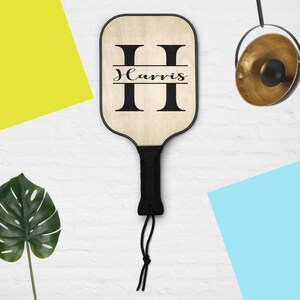 Könnte beinhalten: Ein Pickleball-Schläger aus Holz mit schwarzem Griff und dem Namen "Harris" in schwarzen Buchstaben. Der Schläger hat einen schwarzen Rand und einen schwarzen Griff. Der Schläger hängt an einer weißen Wand.