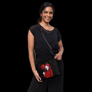May include: Black crossbody bag featuring a red and white illustration of a clown holding a red balloon. The clown is wearing a red hoodie. The bag has a black adjustable strap and a zipper closure. The bag is made of a durable material.