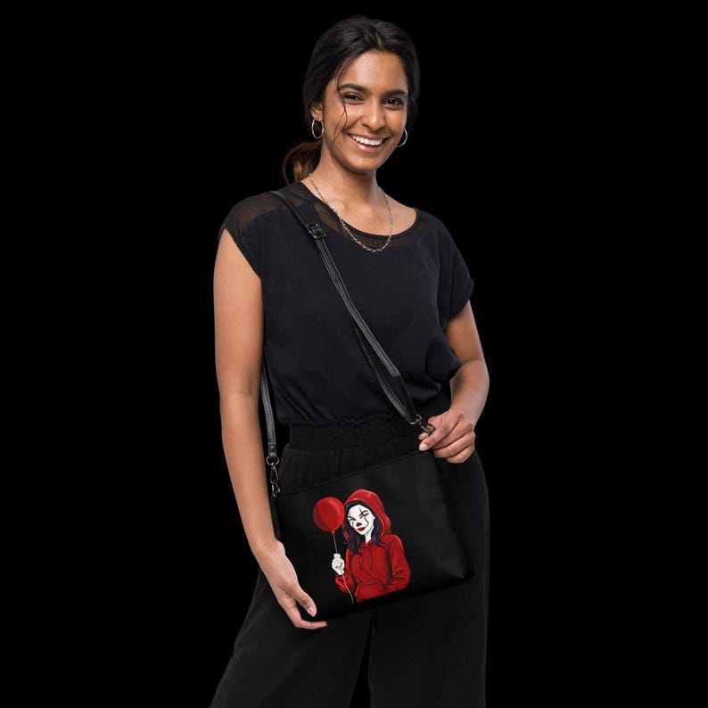May include: Black crossbody bag featuring a red and white illustration of a clown holding a red balloon. The clown is wearing a red hoodie. The bag has a black adjustable strap and a zipper closure. The bag is made of a durable material.