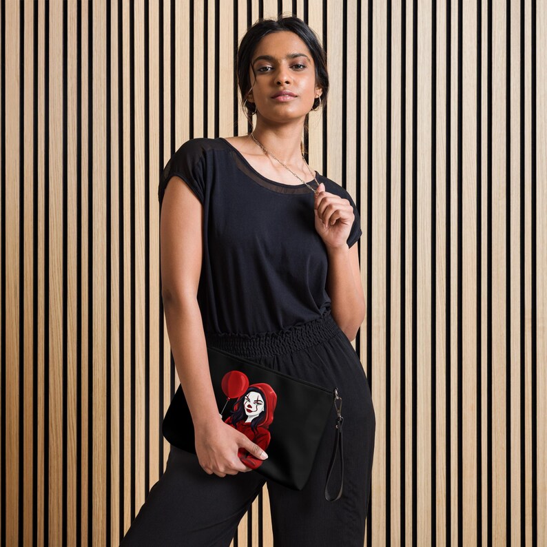 May include: Black clutch featuring a red clown illustration holding a red balloon. The clutch has a wrist strap and a zipper closure. The background is a vertical wood panel design.