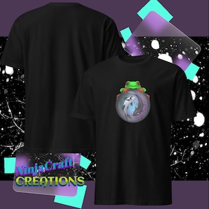 May include: Black t-shirt featuring a colorful graphic of a frog perched atop a circular design with a fish. The design is centered on the front of the shirt. The text "NinjaCraft CREATIONS" is visible in the lower left corner.