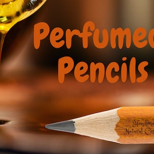 May include: A close-up of a wooden pencil with the text "Handmade Natural Cosmetics" on the side. The pencil is lying on a surface with a golden liquid dripping onto it. The text "Perfumed Pencils" is in the background with the text "La Bouille" in smaller font.