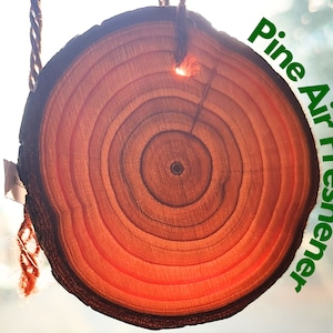 Peut inclure: Une tranche de bois ronde avec des anneaux de croissance visibles, suspendue à une ficelle. La tranche est un désodorisant d'air naturel à l'odeur de pin. Le texte "Pine Air Freshener" est écrit en lettres vertes sur le côté droit de l'image.