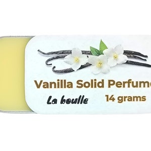 May include: A small, round tin container with a white label that reads "Vanilla Solid Perfume La bouille 14 grams". The label features a graphic of vanilla beans and white flowers.