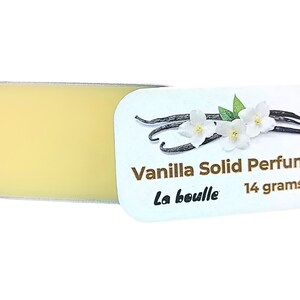 May include: A white tin with a gold label that reads "Vanilla Solid Perfume La bouille 14 grams". The label features a drawing of vanilla beans and white flowers. The tin is open and shows a yellow solid perfume inside.