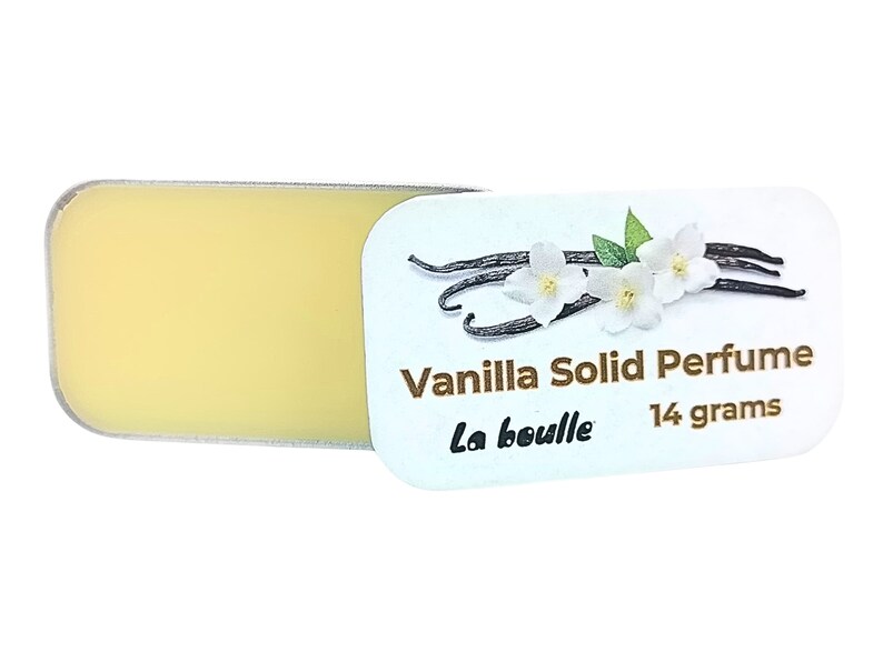 May include: A white tin with a gold label that reads "Vanilla Solid Perfume La bouille 14 grams". The label features a drawing of vanilla beans and white flowers. The tin is open and shows a yellow solid perfume inside.