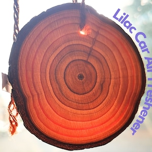 May include: A round wooden car air freshener with a natural wood grain pattern. The air freshener is hanging from a string and has the text "Lilac Car Air Freshener" written on it.