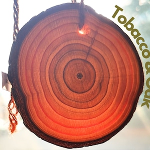 Può includere: Un primo piano di una fetta di legno con anelli visibili, appesa a una corda. Il testo "Tobacco & Oak" è scritto in lettere dorate su uno sfondo sfocato.