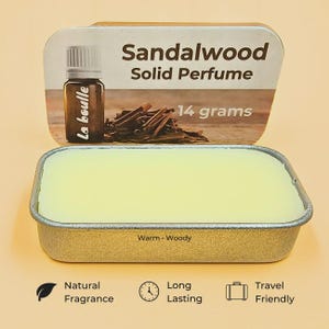 Op de afbeelding: Een rechthoekige blikken doos met vaste sandelhoutparfum en een lichtgele balsem. Op de doos staat "Sandalwood Solid Perfume" en "14 gram". Op de achtergrond een kleine parfumfles en houtsnippers. Het etiket luidt "Warm Woody".