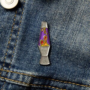 May include: A silver-colored lava lamp pin with a purple and orange lava-like design. The pin is on a blue denim background.