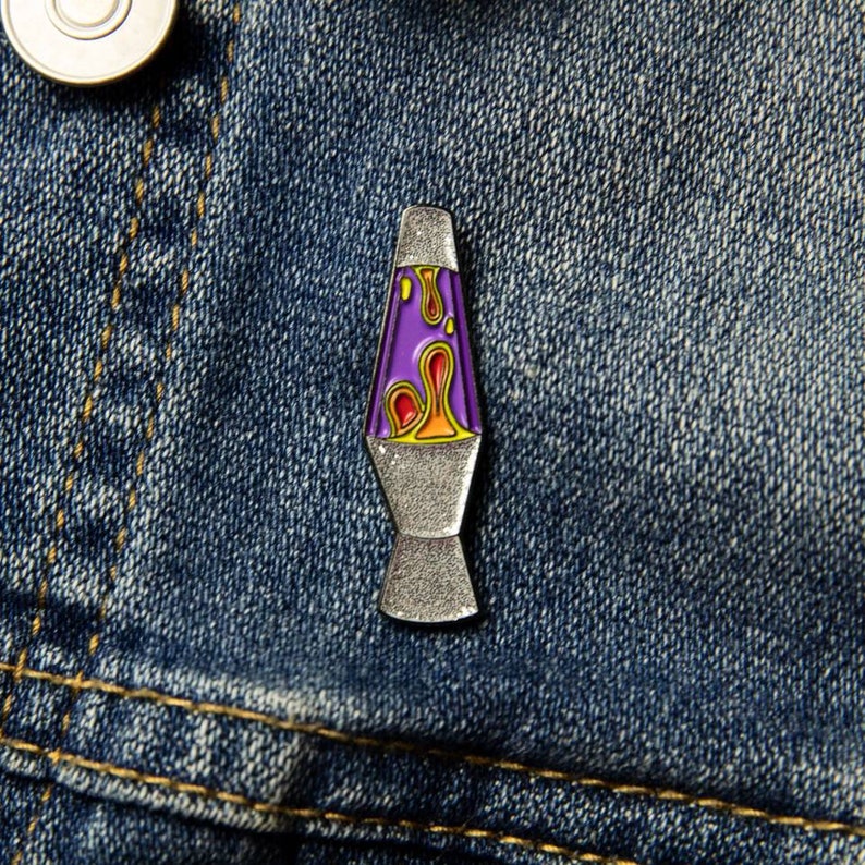 May include: A silver-colored lava lamp pin with a purple and orange lava-like design. The pin is on a blue denim background.