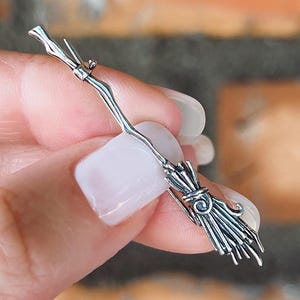 May include: A silver-colored miniature broom charm with detailed bristles and handle. The charm is held in a hand against a blurred brick background. The broom is a Halloween or witch-themed accessory.
