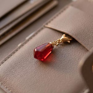 Può includere: Primo piano di un portafoglio in pelle color talpa con un ciondolo rosso a forma di gioiello. Il ciondolo ha la forma di un melograno e pende da una chiusura color oro. Il portafoglio è aperto, rivelando fessure per carte e una chiusura a scatto.