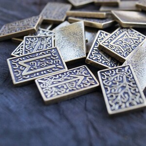 Puede incluir: Un mont&oacute;n de azulejos de lat&oacute;n con dise&ntilde;os y s&iacute;mbolos intrincados. Los azulejos est&aacute;n esparcidos sobre una superficie de madera oscura.