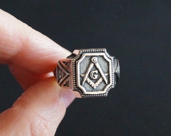 Peut inclure: Une bague en argent avec un motif carré représentant un symbole maçonnique, un compas et une équerre avec un 'G' au centre.
