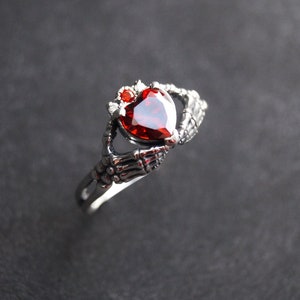 Könnte beinhalten: Ein silberner Ring mit zwei Skeletthänden, die einen roten herzförmigen Edelstein halten.