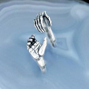 May include: A silver ring featuring two skeletal hands, one grasping the band and the other open. The ring is set against a light blue, marbled background. The ring's design is detailed, showcasing the bones of the hands.