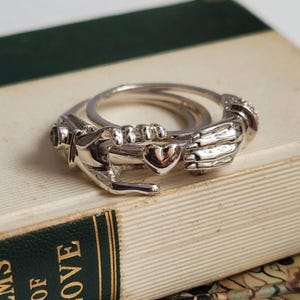 Könnte beinhalten: Zwei silberne Ringe, einer mit Herz und Händen, der andere schlicht. Sie liegen auf einem Stapel alter Bücher, mit dem Titel "POEMS OF LOVE" auf dem Buchrücken. Die Ringe sind detailliert und glänzend.