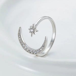 May include: A silver-colored ring featuring a crescent moon design adorned with small, clear stones. A star-shaped element with a central clear stone is positioned opposite the moon, creating an open design. The ring is set against a white background.