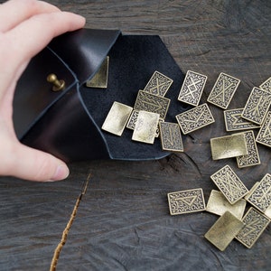 Puede incluir: Una bolsa de cuero negro con un cierre de lat&oacute;n, derramando una colecci&oacute;n de peque&ntilde;as runas rectangulares de lat&oacute;n. Cada runa est&aacute; grabada con un s&iacute;mbolo &uacute;nico.