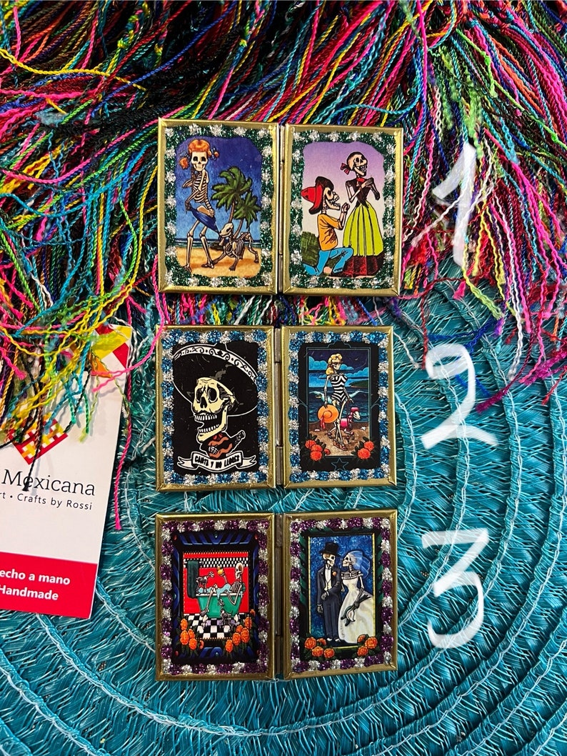 May include: Four small, framed, hinged, decorative boxes with colorful images of skeletons in various scenes. The boxes are decorated with glitter and have gold frames. The boxes are arranged in two rows of two. The boxes are on a blue and green background.