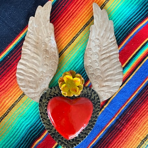 May include: A red heart with gold trim and a yellow flower is surrounded by two brown wings. The heart and wings are on a colorful striped background.