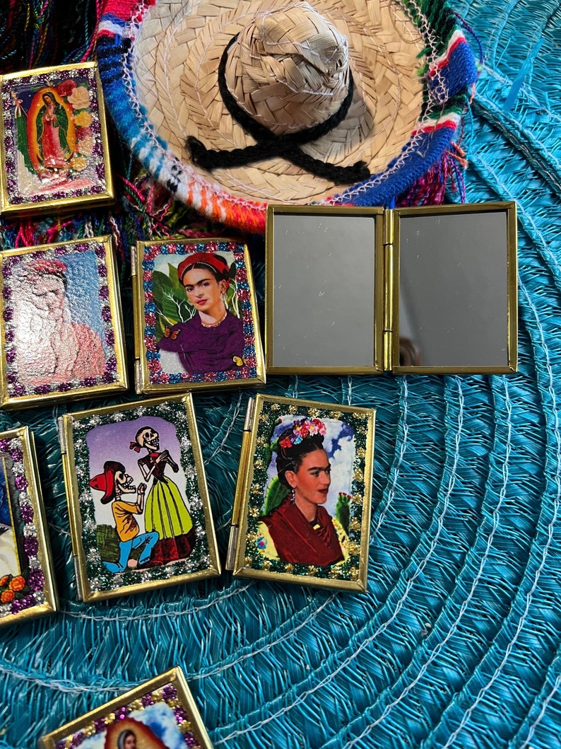 May include: A collection of small, framed, decorative images with gold trim. The images feature various subjects, including religious figures, a woman with flowers in her hair, and a couple dressed as skeletons. The images are arranged on a blue and white patterned surface.