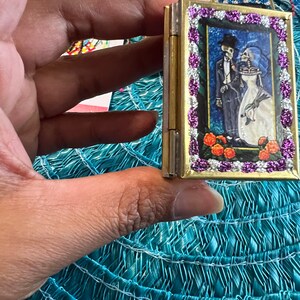 May include: A small, gold-colored, hinged box with a colorful image of a skeleton couple in a wedding setting. The box is decorated with a purple and white beaded border.