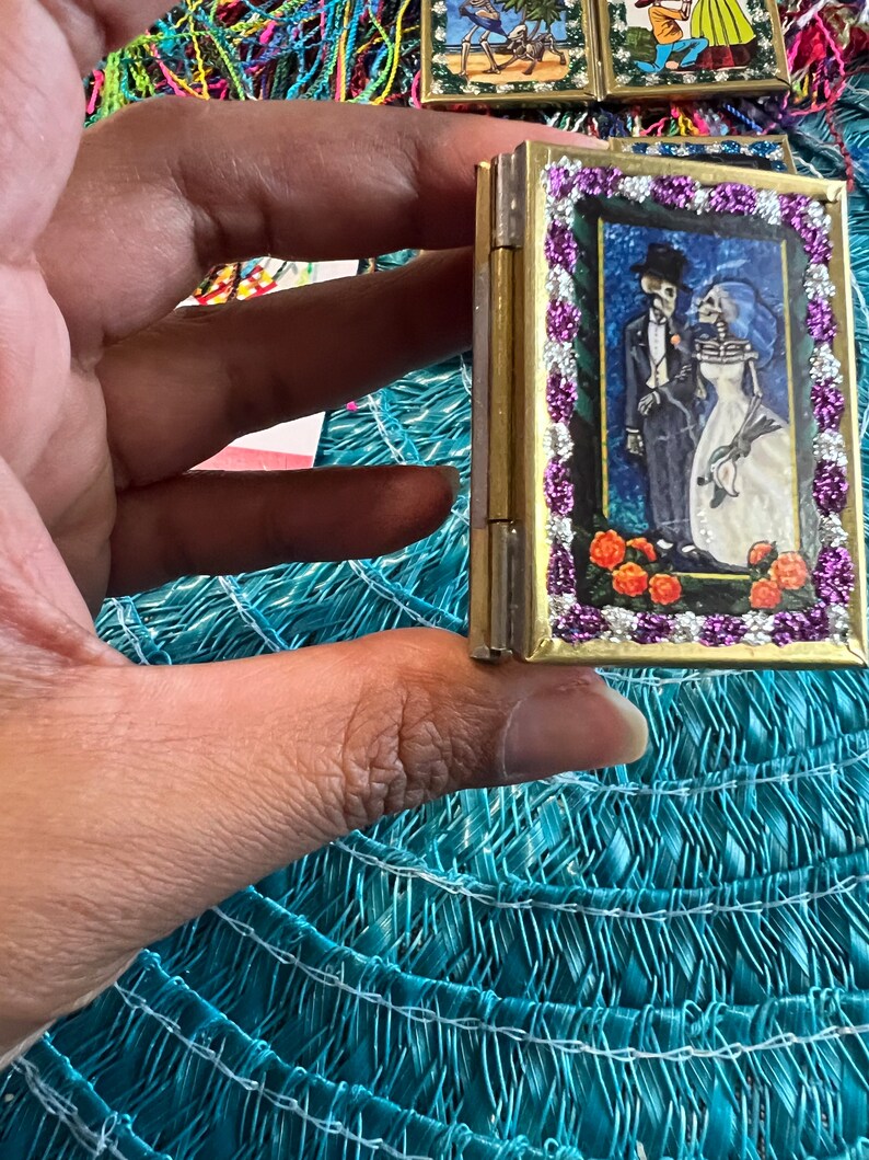 May include: A small, gold-colored, hinged box with a colorful image of a skeleton couple in a wedding setting. The box is decorated with a purple and white beaded border.