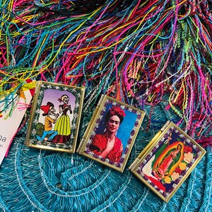 May include: Three small, framed, gold-trimmed pictures with colorful images. The first picture shows a skeleton couple in a romantic setting. The second picture shows Frida Kahlo with a red dress and flowers in her hair. The third picture shows the Virgin Mary with a blue robe and a gold halo.