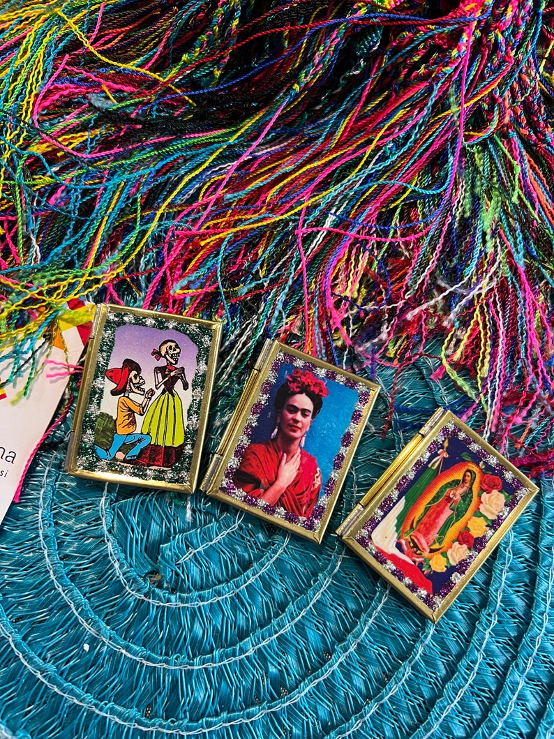 May include: Three small, framed, gold-trimmed pictures with colorful images. The first picture shows a skeleton couple in a romantic setting. The second picture shows Frida Kahlo with a red dress and flowers in her hair. The third picture shows the Virgin Mary with a blue robe and a gold halo.