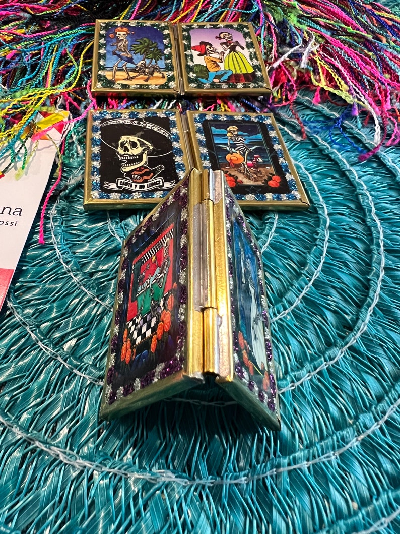 May include: A set of five small, framed, decorative mirrors with gold frames. Each mirror features a different Day of the Dead themed image. The mirrors are arranged in a circle on a blue and green surface.