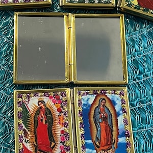 May include: A gold-framed, hinged, folding mirror with two images of the Virgin Mary. The images are surrounded by a gold border with purple glitter. The images are set against a blue sky background with clouds.