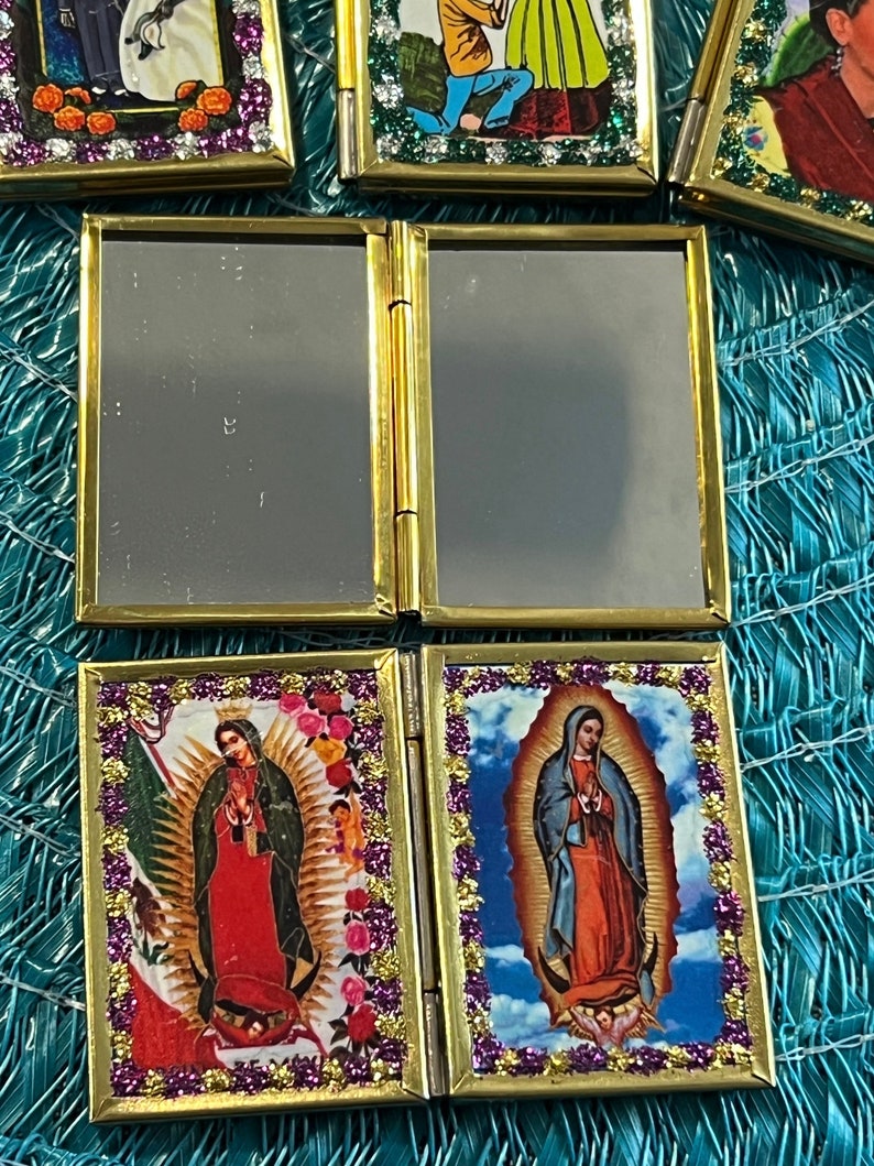 May include: A gold-framed, hinged, folding mirror with two images of the Virgin Mary. The images are surrounded by a gold border with purple glitter. The images are set against a blue sky background with clouds.