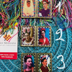 May include: Three small, gold-framed, mirrored picture frames with images of Frida Kahlo. Each frame has a different image of Frida Kahlo. The frames are on a blue and green background with colorful yarn.