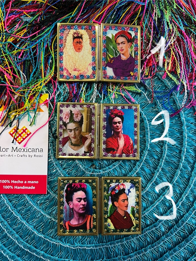May include: Three small, gold-framed, mirrored picture frames with images of Frida Kahlo. Each frame has a different image of Frida Kahlo. The frames are on a blue and green background with colorful yarn.