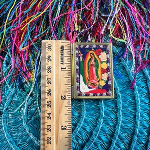 May include: A small, gold-framed picture of the Virgin Mary with a blue robe and a crown of stars. The picture is surrounded by a border of white and gold glitter. The picture is sitting on a blue and green woven mat with a wooden ruler next to it.