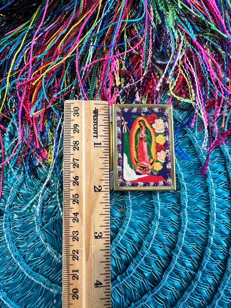 May include: A small, gold-framed picture of the Virgin Mary with a blue robe and a crown of stars. The picture is surrounded by a border of white and gold glitter. The picture is sitting on a blue and green woven mat with a wooden ruler next to it.