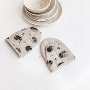 May include: Two beige oven mitts with a hedgehog pattern. The mitts are on a white surface with a stack of beige ceramic bowls and a vase with dried flowers.