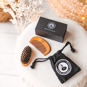 Puede incluir: Un kit de aseo para barba con un peine de madera, un cepillo y una bolsa negra con cordón. El kit incluye una caja negra con el texto "Beard Brush & Comb Set". Los artículos están dispuestos sobre una superficie blanca.
