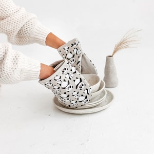 Puede incluir: Un par de guantes para horno con un diseño de estampado de panda. Los guantes son de color beige con caras de panda negras. Los guantes están colocados encima de una pila de platos de cerámica.