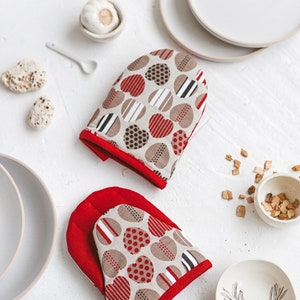 Puede incluir: Un par de guantes para horno rojos y beige con un patrón de corazón. Los guantes están hechos de tela y tienen un ribete rojo.