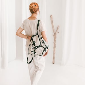 Può includere: Una donna indossa una blusa beige chiaro e pantaloni bianchi e porta uno zaino beige e nero con un motivo floreale. Lo zaino ha una chiusura a coulisse e una patta con una fibbia.