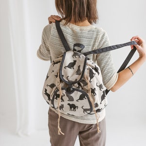 May include: A beige canvas backpack with black cat silhouettes printed all over. The backpack has a drawstring closure and adjustable straps.