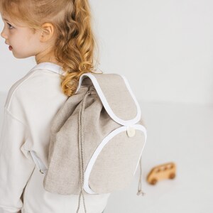 May include: A young girl wearing a white sweatshirt and blue jeans is wearing a beige linen backpack with white trim. The backpack has a drawstring closure and a single button.