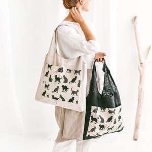 May include: Two tote bags, one beige with black cat silhouettes and one black with white cat silhouettes. Both bags have a pocket on the front.