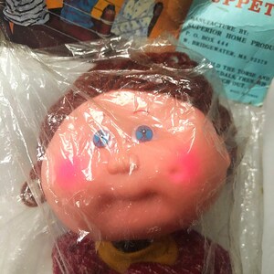 Puede incluir: Una muñeca Punch Puppet vintage en una bolsa de plástico. La muñeca tiene una cara color melocotón con mejillas rosadas, ojos azules y cabello castaño. Lleva un suéter burdeos con una pajarita amarilla. El embalaje incluye el texto "Punch Puppet".