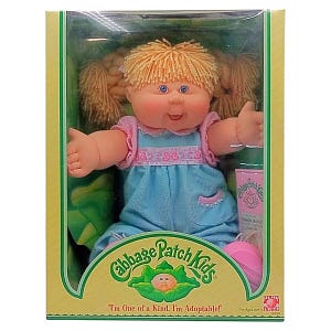 May include: A Cabbage Patch Kids doll in its original packaging. The doll has blonde pigtails, blue eyes, and is wearing a blue dress with pink trim. The packaging is green and yellow, with the Cabbage Patch Kids logo and the text "I'm One of a Kind. I'm Adoptable!"